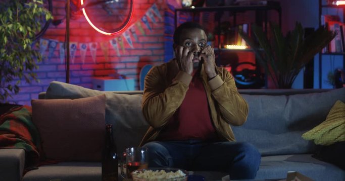 African American young guy sitting on the couch in the dark room at night and watching scary movie on TV.