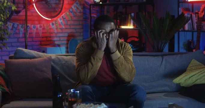 Portrait shot of the young African American man sitting on the sofa in the living room and watching horror movie on TV at night.