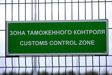 Customs control zone - a sign in Russian and English at the entrance to the vehicle inspection point. The plate is green on the metal grill. Pointer and warning about entering  special area.