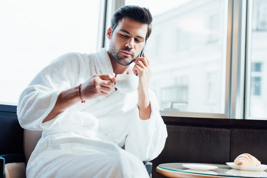  bearded man in white bathrobe holding cup and talking on smartphone
