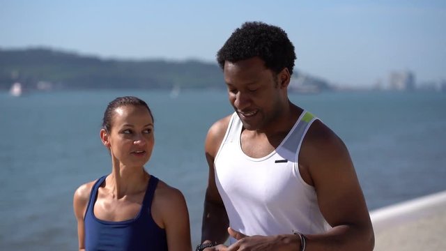 Multiethnic couple in sportswear jogging along embankment. Happy sporty young couple running together and talking at riverside at sunny day, slow motion. Workout concept
