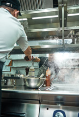 Help in kitchen. Vertical photo of young professional chef giving a pepper grinder to his assistant while cooking process.