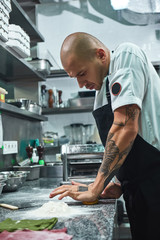 Working process. Vertical photo of handsome professional chef with tattoos on his hands kneading the dough in restaurant kitchen