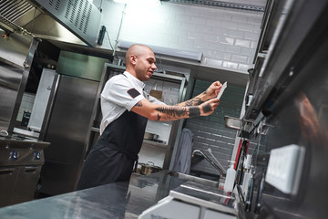 Fototapeta na wymiar What is next... Handsome bald male chef with tattoos on his arms looking at order list in a restaurant kitchen.