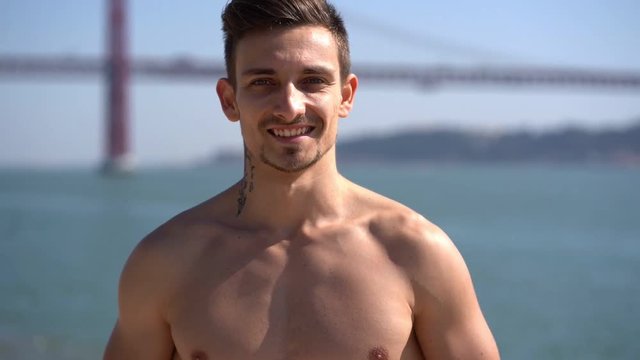 Handsome bare-chested sportsman smiling at camera outdoor. Portrait of muscular shirtless man standing near river and looking at camera, handheld shot. Healthy lifestyle concept