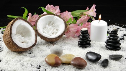 Two halves of a coconut , stones, rocking balancing and a burning candle are on a wooden table covered with coconut chips, against a background of pink flowers