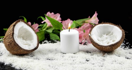 Obraz na płótnie Canvas Two halves of a coconut and a burning candle are on a wooden table covered with coconut flakes, against a background of pink flowers