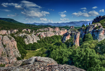 Dynamic contrast landscape of a rocky valley with green vegetation, surrounded by natural sculptures made of rock, distant mountain ridge on the horizon, blue sky with a few clouds