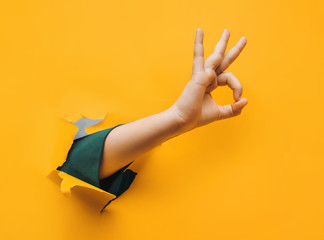 Hand OK sign. The teenage hand came out into the hole of yellow paper and shows symbol of Okay. Copy space, fine. - obrazy, fototapety, plakaty