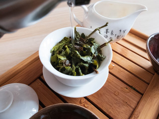 Pouring a boiled water to tea brewing vessel with leaves of oolong tea on tea draining tray.