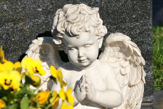 Betender Friedhofsengel auf einem Grab mit unscharfen Frühlingsblumen im Vordergrund