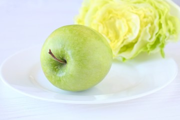 Fresh organic green juicy apple on white plate with half of green lettuce salad on wooden neutral background. Diet concept with raw vegetarian food. Green fruit and vegetables at the dish