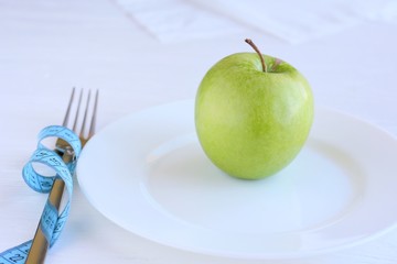Fresh organic green juicy apple on white plate with blurred fork and knife and blue measuring tape on wooden neutral background. Diet concept with raw vegetarian food. Green fruit at the dish