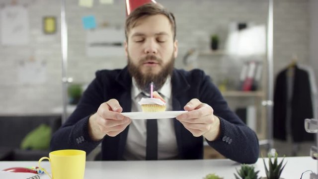 Sad bearded businessman, office worker is celebrating a lonely birthday in the office, he is blowing a candle on a small cake. He wears an office suit, white shirt and blak tie.