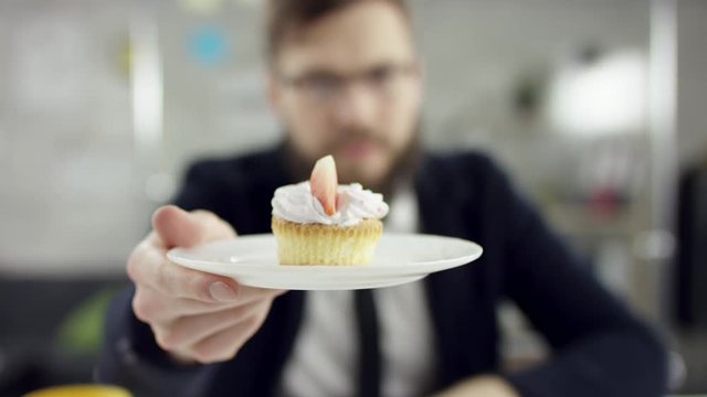Sad working bearded businessman, office worker is celebrating a lonely birthday in the office, he is showing and giving a cup-cake. He wears an office suit, white shirt and blak tie.