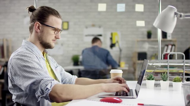 young man singing sitting at office desk with laptop surrounded by diverse colleagues, lazy worker wasting time at workplace in co-working. he is drinking coffee. Side view