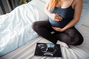 Pregnant woman sitting on bed and looking at ultrasound photo of her baby