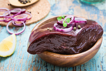 A piece of raw beef liver in a bowl, onion, lemon and spices for cooking on a wooden table