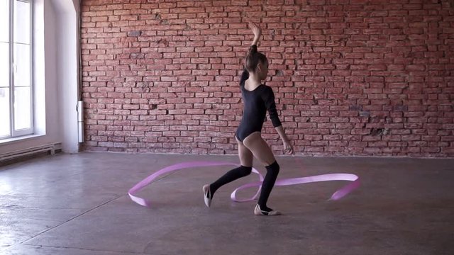 Rhythmic gymnastics for girls. Cute girl exercising in dancing stuido alone.Dancing, spinning with pink ribbon. Slow motion footage. Brick wall on the background