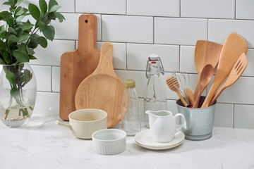 Simple rustic kitchenware against white wooden wall: rough ceramic pot with wooden cooking utensil set, stacks of ceramic bowls, jug and wooden trays.