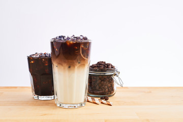 Iced coffee and latte macchiato in tall glass with straws and coffee beans in glass jar on wooden table over white background. Various types of coffee. Copy space for text.