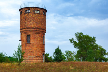 Red brick water tower on a hill against the backdrop of a rural landscape. A single-standing building with a tank for water supply.