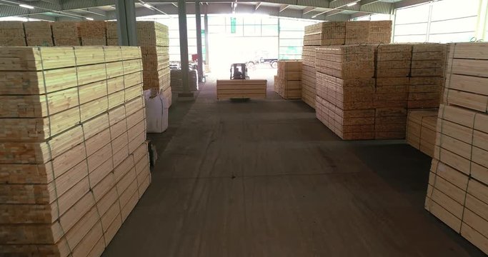 A large warehouse of timber materials. A forklift carries a wooden beam through the warehouse. Neatly stacked wooden bar in a sawmill warehouse