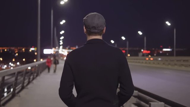 A young elegant man walks on the sidewalk along the motorway in the evening