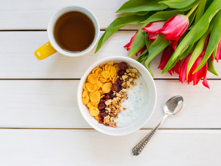 Delicious yogurt bowl with corn flakes, nuts and jam on a white wooden table. Healthy and organic nutrition concept. Tulips with cup of tea and breakfast. Top view, copy space