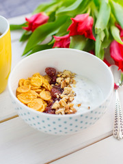 Delicious yogurt bowl with corn flakes, nuts and jam on a white wooden table. Healthy and organic nutrition concept. Tulips with cup of tea and breakfast.