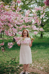 Fototapeta na wymiar beautiful cute brunette girl bride in cream lace dress, with decoration on the hair, near the tree blossoms with pink flowers, garden, greenwich park