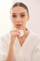 Thoughtful young woman in bathrobe standing with cotton pad