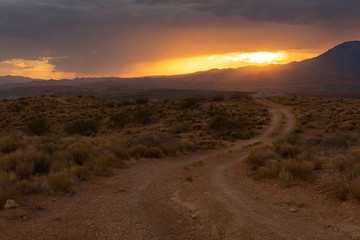 Fototapeta na wymiar Sunset light glows orange over a landscape with a dirt road leading into the desert with the sun going down behind clouds and a distant mountain.