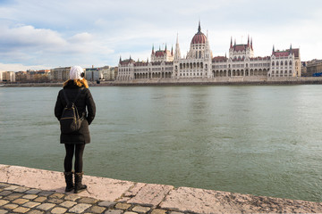 Young woman is standing on the riverbank of the Danube river and looking at Hungarian Parliament Building in BudaPest, Hungary
