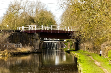 Fototapeta premium The London-Rugby railway bridge crosses over the Grand Union Canal in Whilton Locks. Landscape looking up the waterway, with water cascading through the closed lock gates. Northamptonshire. England.