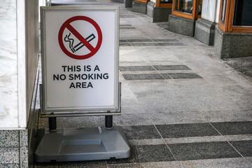  This is a no smoking area sign on posh looking street, marble pavement, and bottom walls of shops.