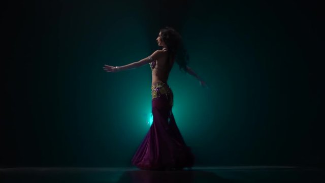 Girl performs in a dark studio belly dance in oriental costume. Smoke background. Slow motion