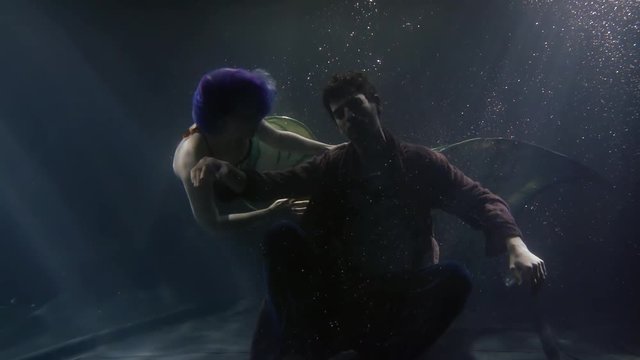 mermaid is swimming around drowned man near ground of sea in darkness, underwater