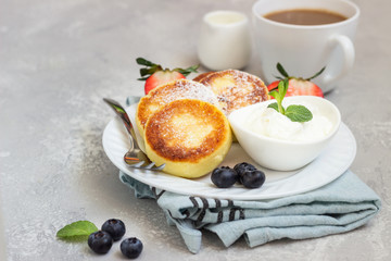 Cottage cheese fritters (syrniki) served with sour cream, fresh berries (strawberry and blueberry) and mint on a light grey background. Healthy breakfast or diet lunch. Copy space.