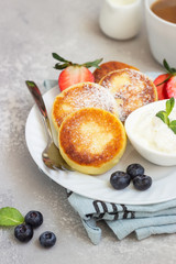 Cottage cheese fritters (syrniki) served with sour cream, fresh berries (strawberry and blueberry) and mint on a light grey background. Healthy breakfast or diet lunch. Copy space.