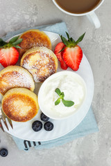 Cottage cheese fritters (syrniki) served with sour cream, fresh berries (strawberry and blueberry) and mint on a light grey background. Healthy breakfast or diet lunch. Copy space.