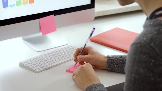 Female hands with red nails write note on pink post it and put it on the computer next to the white keyboard