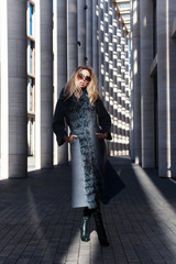 Stylish woman in rich fur coat and sunglasses in modern city street columns background, autumn fashion trend, green boots, urban style, accessories, legs, footwear. Autumn or spring fashion shopping
