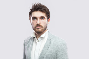 Portrait of handsome bearded serious man with brown hairs and beard in white shirt and casual grey suit standing and looking at camera. indoor studio shot, isolated on light grey background.