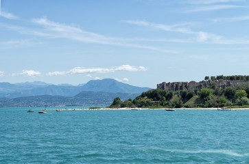 Fototapeta premium Stony Beach of Sirmione town on Garda Lake with view of Grottoes of Catullus (Grotte di Catullo), the ruins of a Roman villa built at the end of the 1st century B.C. View from Garda Lake.