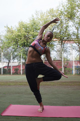 young skinhead woman standing in yoga tree pose near green trees in summer park