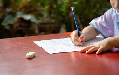 little Thai student in uniform school suit hold blue pencil writing her homework near garden. practice writing on red wooden table. Alphabet tracing worksheet A to Z.
