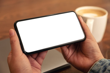 Mockup image of hands holding black mobile phone with blank desktop screen horizontally with coffee cup on the table