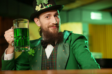 Dark-haired bearded young man in a leprechaun hat with shamrock raising his glass with green beer