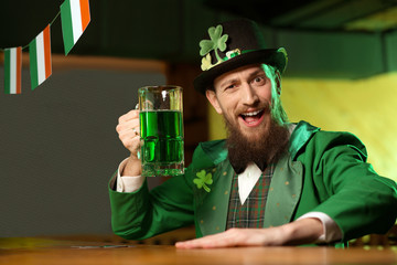 Dark-haired bearded young man in a leprechaun hat with shamrock drinking green beer